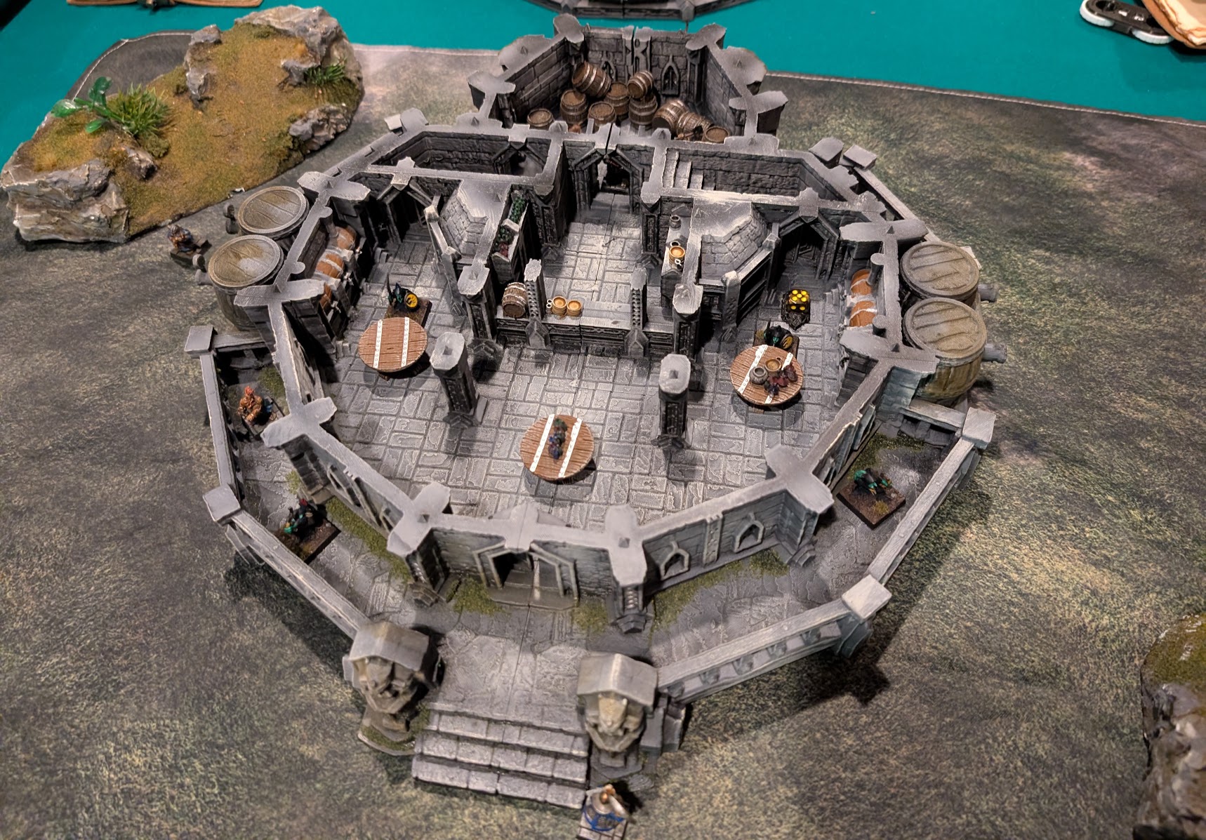 Miniatures occupy a roofless circular tavern with a central hearth, tables, benches, and barrels.
